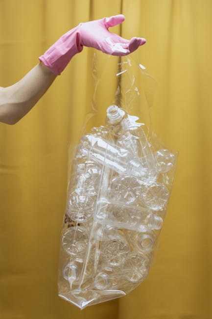 A person wearing a pink rubber glove is holding a clear, crumpled plastic bag filled with various empty plastic bottles and containers. The bottles exhibit different shapes, sizes, and textures, mostly transparent with some showing slight sheen or reflections of light. The background is a plain yellow curtain or wall, creating a neutral environment suitable for illustrating waste collection or rubbish clearance practices. The scene emphasizes the handling of plastic waste, which is relevant to private rubbish removal services such as those provided by Rubbish Clearance Kensington, supporting the context of alternative waste management and on-site clearance procedures.
