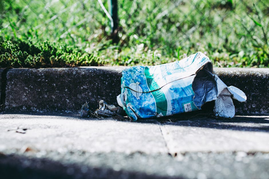 A crumpled plastic water bottle wrapper lies on the concrete pavement next to a small curb, with the wrapper featuring blue, green, and white colors and an image of a fish. The wrapper is partially torn and flattened, with some of its edges curled. Behind the curb, there is a patch of green grass with varying shades and a slightly blurred background of foliage, indicating an outdoor urban or suburban environment. The sunlight casts shadows on the pavement, highlighting the textures of the concrete surface and the plastic waste. This scene exemplifies roadside litter, which may be addressed through private waste collection services offered by Rubbish Clearance Kensington, as part of efforts to maintain cleaner streets and reduce environmental impact from improper rubbish disposal.