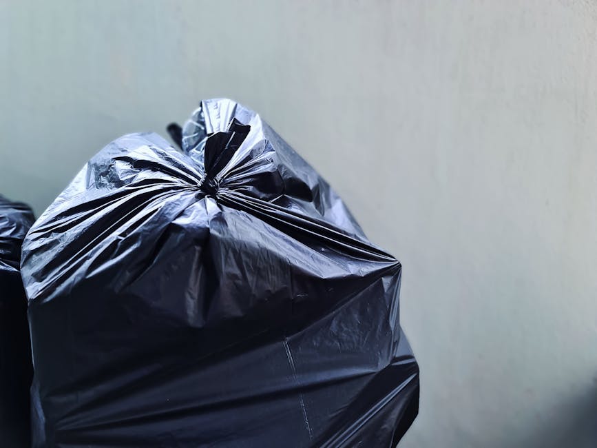 A close-up of a black plastic rubbish bag tightly tied at the top, resting against a plain, light-colored wall. The bag is made of glossy, crinkled plastic material, with visible creases and folds, indicating it is filled with waste. The surrounding environment appears to be an indoor or sheltered area, with no other objects visible nearby, suggesting it may be part of a collection or disposal process managed by a private waste removal service such as Rubbish Clearance Kensington. The lighting is natural or diffuse, highlighting the shiny surface of the bag and emphasizing its bulging shape, which reflects typical rubbish collection practices where waste is prepared for removal. This scene illustrates typical waste storage prior to independent rubbish collection or disposal, relevant to alternative waste handling methods outside standard municipal services, aligning with concepts discussed in the page about common problems with rubbish collection on Kensington mews.