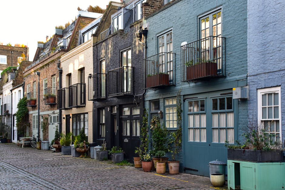 A narrow cobblestone alleyway lined with potted plants and greenery along a low brick wall on the left side, with residential terraced houses on the right, featuring white-framed sash windows, brick and white-painted facades, and black drainage pipes attached to the walls. A large tree with dense green foliage extends overhead, partially obscuring the partly cloudy sky. The scene appears quiet and well-maintained, reflecting an urban residential environment, which may occasionally require private or alternative waste clearance services. The absence of visible rubbish or clutter suggests the area is tidy, but the proximity of potted plants and the narrow nature of the alley highlight the importance of effective rubbish collection for such communal outdoor spaces. Rubbish Clearance Kensington offers collection solutions that could be suitable for managing waste in these settings, supporting proper waste handling in urban residential mews. Lighting is natural with soft daylight, and shadows cast by the tree and buildings emphasize the vertical and horizontal textures of the brickwork and pavement, contributing to a calm, orderly atmosphere typical of such private alleyways.