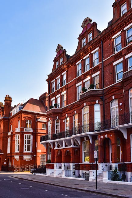 The image depicts a row of historic red-brick Victorian-style residential buildings situated along a residential street, with the focus on a multi-story building featuring ornate architectural details such as decorative cornices, arched windows, and wrought iron balconies. The facade exhibits a mixture of deep red brickwork with contrasting white accents around the windows and balcony supports, creating a visually textured appearance. Large windows on each level reflect daylight, and some windows have potted plants on the balcony railings. The buildings are set back slightly from the pavement, which is lined with a low metal railing and street furniture, including a parking sign. The street appears quiet, with minimal vehicular presence, and the natural daylight highlights the rich tones of the brickwork against a clear sky. This urban scene subtly reflects the context of private or alternative waste management options, suggesting a well-maintained residential setting where rubbish clearance services such as those provided by Rubbish Clearance Kensington may operate for efficient waste removal, distinct from council collection routines.