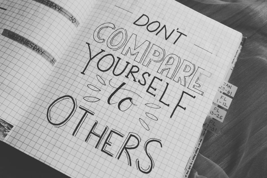 A close-up view of a spiral-bound notebook with grid paper. On the page, there is a handwritten message reading 'DON'T COMPARE YOURSELF to OTHERS' in large, bold capital letters, with the word 'yourself' emphasized in a slightly different style. The message is surrounded by small decorative lines and swoops, drawn with a black pen. In the background, partly visible, is a wooden desk or table surface with some loose paper or sticky notes attached to the edge of the notebook. The overall lighting is soft, and the scene appears to be indoors, suggesting an environment where someone is journaling or reflecting. Although the message is motivational, it subtly connects to themes of personal growth and individual effort, which can align with private or independent approaches to responsibilities like rubbish removal, managed by services such as Rubbish Clearance Kensington.