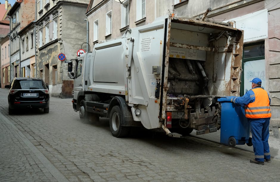 A municipal waste collection truck, painted white with visible rust and wear on its rear loading area, is parked on a narrow cobbled street lined with multi-story residential buildings with aged, textured facades. The truck's rear is open, showing mechanical components and a partially loaded compartment waiting for rubbish disposal. A worker wearing a high-visibility orange vest with blue overalls and a blue cap is positioned on the right, actively handling a blue wheeled bin. The worker’s stance indicates they are either placing the bin on a designated collection area or preparing it for emptying. To the left of the truck, a black parked car is positioned close to the curb, and behind the vehicle, there is a no parking or stopping sign mounted on a pole attached to one of the residential buildings. The scene suggests a typical urban rubbish collection operation, supporting alternative waste handling methods outside of official communal refuse schemes, with a focus on private collection or on-site clearance activities as managed by waste management services such as Rubbish Clearance Kensington.