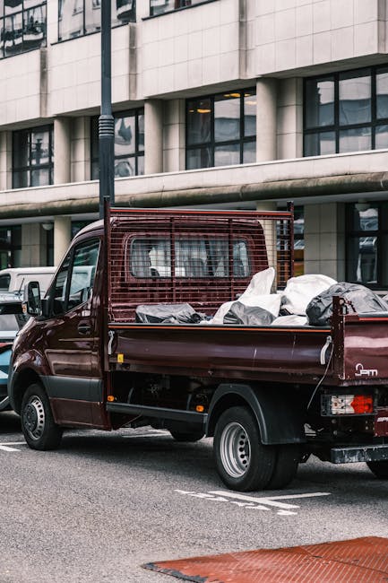 A small dark red flatbed truck parked on a city street with its cargo bed loaded with various types of rubbish, including black plastic bags, white wrapped items, and debris. The truck features a metal cage guard behind the cab for securing cargo and is positioned next to a modern building with large glass windows and a stone facade. The vehicle is in a designated parking zone, with other parked cars visible in the background. The scene is illuminated by natural daylight, and the environment suggests an urban setting suitable for private waste collection services such as those offered by Rubbish Clearance Kensington, which may perform alternative on-site clearance or rubbish removal tasks in areas like Holland Park estate, aligning with local rubbish management rules.