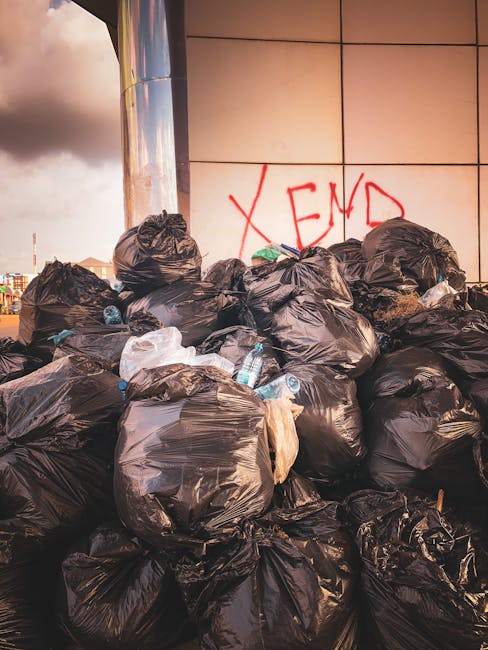 A large accumulation of black plastic rubbish bags piled against a modern building with a tiled facade, some of which are torn open revealing discarded plastic bottles and other waste inside. The bags are haphazardly stacked in the foreground, with a few loose items such as bottles and small pieces of trash scattered around their base. Behind the piles, there is a wall with graffiti in red spray paint spelling 'XEND' and a partly cloudy sky is visible in the background, indicating an outdoor urban environment. The scene depicts an example of illegal or unmanaged rubbish accumulation that could fall under private waste disposal or alternative rubbish clearance services provided by companies like Rubbish Clearance Kensington, illustrating the need for professional rubbish removal to responsibly manage waste alongside preventing fly tipping and ensuring compliance with local disposal regulations.
