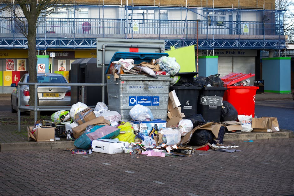 A large open-top metal bin designated for mixed paper and cardboard collection is positioned on a paved urban sidewalk, surrounded by numerous overflowing rubbish bags made of black plastic and brown paper. The waste includes crumpled cardboard boxes, loose newspapers, plastic packaging, and discarded appliances, some partially flattened or torn. Next to the bin, there are several loose piles of waste spilling onto the ground, with additional cardboard boxes and packaging materials scattered nearby. To the left of the waste, a silver car with a visible license plate is parked adjacent to a metal railing, which separates the rubbish area from the rest of the sidewalk. In the background, a commercial building with a blue construction scaffold partially obscures store entrances and signage, indicating ongoing refurbishment. The scene is illuminated by natural daylight, highlighting the cluttered state of the refuse collection point, typical of private waste handling arrangements that may temporarily exceed local authority collection capacity, making use of private rubbish clearance services such as Rubbish Clearance Kensington.