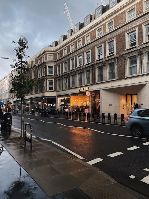 A multi-storey building with a brick and neutral-toned facade, featuring large rectangular windows on each floor, located on a city street. The ground floor houses a retail shop with a brightly lit interior, displaying a yellow-colored sign that reads 'JANE BOUL', and large display windows showing products and interior furnishings. The building occupies the right side of the image, with a wide sidewalk in front that appears wet, possibly after rain, reflecting the surrounding environment. Several bollards line the edge of the pavement, separating it from the street, which has a single lane with a dashed white line in the middle, and a moving dark grey SUV visible on the right. In the background, a construction crane extends above the building, indicating ongoing development nearby. To the left, a tree with sparse foliage is situated on the sidewalk, and a few pedestrians are seen near the storefront. This scene exemplifies urban commercial activity, where private waste management providers like Rubbish Clearance Kensington might be involved in alternative rubbish removal services for retail premises in the area.