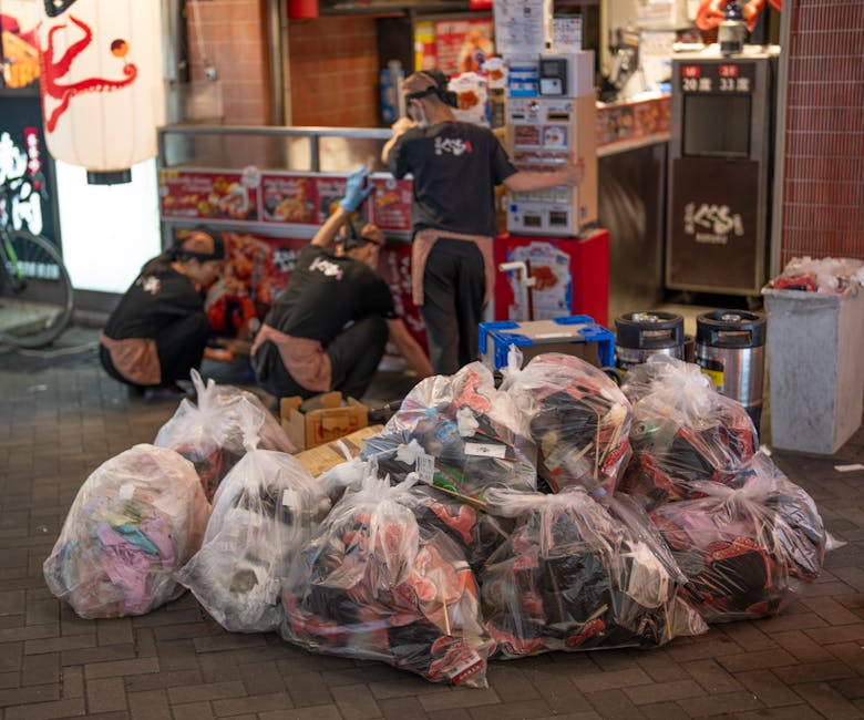 A large open-top metal bin designated for mixed paper and cardboard collection is positioned on a paved urban sidewalk, surrounded by numerous overflowing rubbish bags made of black plastic and brown paper. The waste includes crumpled cardboard boxes, loose newspapers, plastic packaging, and discarded appliances, some partially flattened or torn. Next to the bin, there are several loose piles of waste spilling onto the ground, with additional cardboard boxes and packaging materials scattered nearby. To the left of the waste, a silver car with a visible license plate is parked adjacent to a metal railing, which separates the rubbish area from the rest of the sidewalk. In the background, a commercial building with a blue construction scaffold partially obscures store entrances and signage, indicating ongoing refurbishment. The scene is illuminated by natural daylight, highlighting the cluttered state of the refuse collection point, typical of private waste handling arrangements that may temporarily exceed local authority collection capacity, making use of private rubbish clearance services such as Rubbish Clearance Kensington.