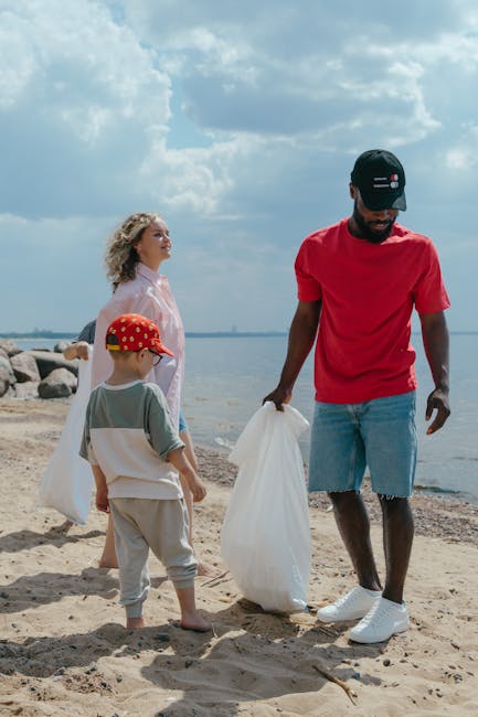 A man and two children are on a sandy beach near the water, involved in a cleanup activity. The man, dressed in a red t-shirt, denim shorts, and a black cap, is holding a large white plastic bag, appearing to collect litter. One child, wearing a red hat with a pattern, a beige and white outfit, is standing nearby and facing the man, observing the activity. The other child, a girl with curly blonde hair, dressed in a light pink outfit, stands slightly behind the boy, looking towards the shoreline. Rocks are visible on the left side of the image, with the calm water stretching into the distance under a partly cloudy sky. The scene depicts an outdoor environment focused on private waste handling, aligning with the theme of independent rubbish collection, while [COMPANY_NAME] could facilitate such community or personal rubbish clearance efforts. The lighting appears natural, with daylight illuminating the scene clearly, emphasizing the textures of the sand, clothing, and plastic bags, reflecting a community-oriented effort to maintain a clean environment outside urban or public waste disposal settings.