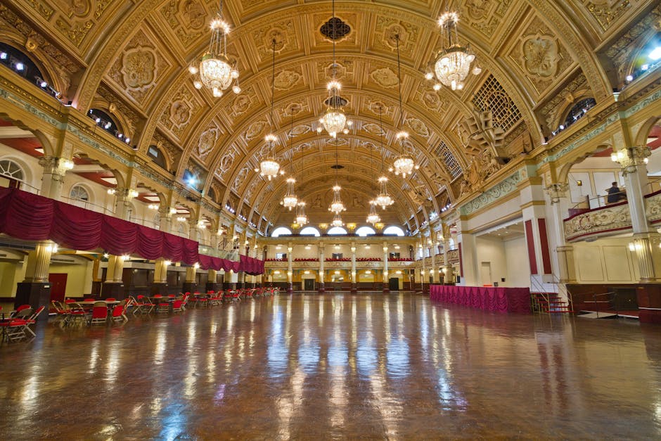 Inside a grand, historic hall with a high, ornate gilded ceiling featuring intricate patterns and multiple decorative chandeliers hanging from above. The spacious wooden floor is polished and reflective, extending across the entire area. Along the sides, there are rows of red chairs and small tables, with deep red velvet drapes hanging from the upper balcony level, which is supported by white columns with gold accents. The lighting is bright, highlighting the elegant details of the architecture, including arched windows at the top allowing natural light to filter in. The overall scene appears clean and well-maintained, suitable for hosting events such as performances or receptions. This interior setting provides a contrast to typical rubbish removal environments but illustrates the importance of professional waste management in maintaining such spaces, possibly through private disposal or on-site clearance services. The scene’s grandeur and meticulous finishing touches emphasize the value of careful rubbish removal planning for large, prestigious venues.
