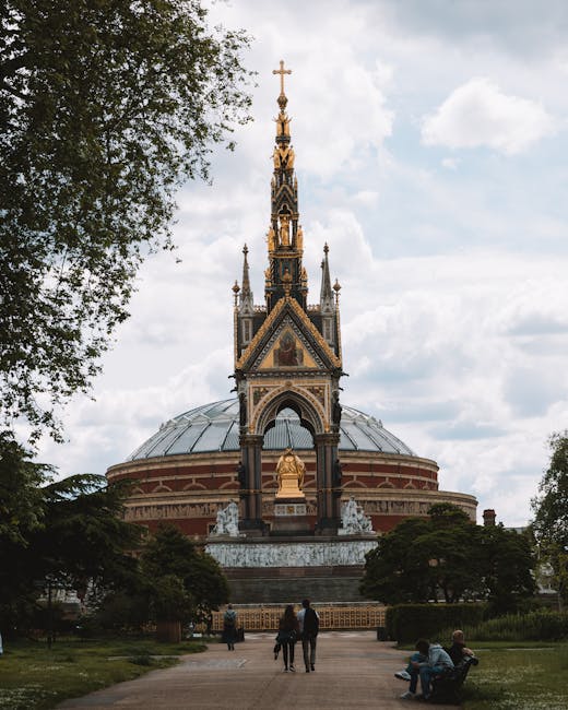 The image depicts a historical monument with a large, domed roof made of metal, set in a landscaped park area. The monument features Gothic Revival architectural elements, including a tall, ornate spire decorated with intricate patterns and topped with a cross. The structure's façade incorporates golden accents, multiple small statues, and detailed carvings. The monument is primarily constructed from stone, with finishes that include polished surfaces and textured details. Surrounding the monument are neatly maintained bushes, trees, and grassy areas, with a paved pathway leading up to the entrance. In the foreground, three people are walking toward the monument, with two individuals standing and observing, and a person sitting on a bench to the right, all dressed casually. The scene is under natural daylight, with a partly cloudy sky providing soft, diffused light. The setting resembles an urban park, where the monument acts as a central feature, and the scene subtly aligns with alternative waste management or private clearance activities, as the area appears clean and well-maintained, possibly indicating on-site rubbish removal or landscaped clearance conducted by professionals such as Rubbish Clearance Kensington.