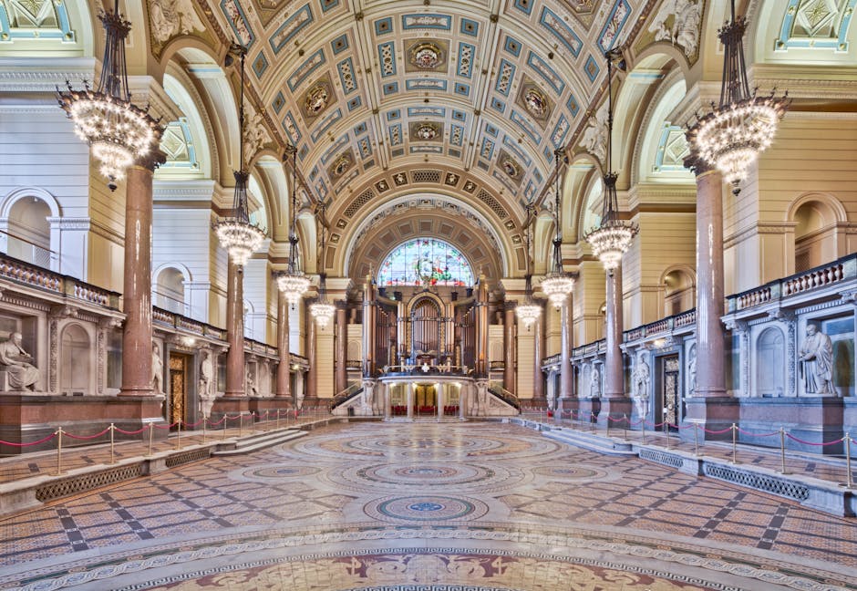 Inside a grand, historic hall with a high, ornate gilded ceiling featuring intricate patterns and multiple decorative chandeliers hanging from above. The spacious wooden floor is polished and reflective, extending across the entire area. Along the sides, there are rows of red chairs and small tables, with deep red velvet drapes hanging from the upper balcony level, which is supported by white columns with gold accents. The lighting is bright, highlighting the elegant details of the architecture, including arched windows at the top allowing natural light to filter in. The overall scene appears clean and well-maintained, suitable for hosting events such as performances or receptions. This interior setting provides a contrast to typical rubbish removal environments but illustrates the importance of professional waste management in maintaining such spaces, possibly through private disposal or on-site clearance services. The scene’s grandeur and meticulous finishing touches emphasize the value of careful rubbish removal planning for large, prestigious venues.