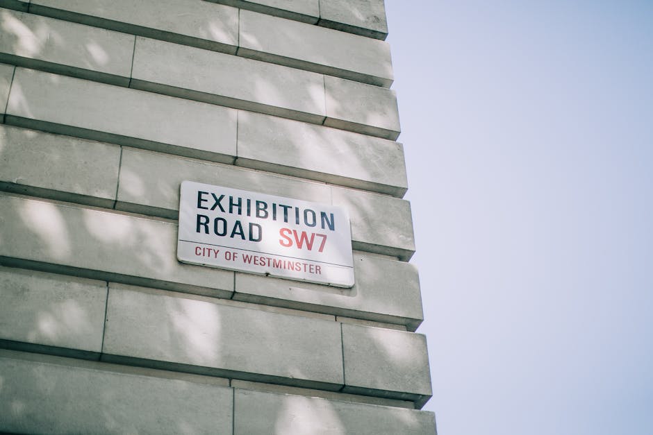 A close-up view of a white rectangular street sign attached to a building's grey stone facade in the City of Westminster, displaying the text 'Exhibition Road SW7' with 'SW7' in red, and 'City of Westminster' in smaller red letters below. The building's exterior features horizontal stone blocks with a smooth finish, casting subtle shadows due to natural daylight. The sky in the background is clear and bright blue, with a few faint shadows of tree leaves partially visible on the wall, indicating nearby greenery. The sign's placement on the wall suggests an urban environment, typical of areas with established architecture and street signage, which may be relevant in the context of implementing regulations for rubbish removal or private waste collection on designated addresses within the district. Rubbish Clearance Kensington provides services related to such private waste handling, supporting alternative disposal solutions beyond local authority channels, especially for projects requiring on-site clearance or independent collection options.