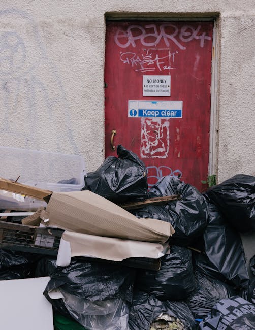 A large pile of black rubbish bags and discarded flattened cardboard boxes stacked against a textured, pale beige wall with visible marks and patches. The bags are tightly tied and appear to contain general waste, with some resting on top of discarded furniture or packaging materials. Behind the pile, there is a red metal door set into the wall, which features various graffiti tags and stickers, including a white sign that reads 'No Money' and a blue and white label that says 'Keep Clear.' The scene is outdoors, situated on a concrete or paved surface, suggesting a back alley or service area. The lighting is natural, with diffused light illuminating the scene evenly. The image subtly relates to private waste disposal practices in urban areas, with the accumulated rubbish indicating the need for scheduled rubbish removal services, such as those provided by Rubbish Clearance Kensington, especially where local authority rules for skip permits are not followed. Overall, it highlights the importance of proper waste management and alternative collection options in busy residential or commercial settings.