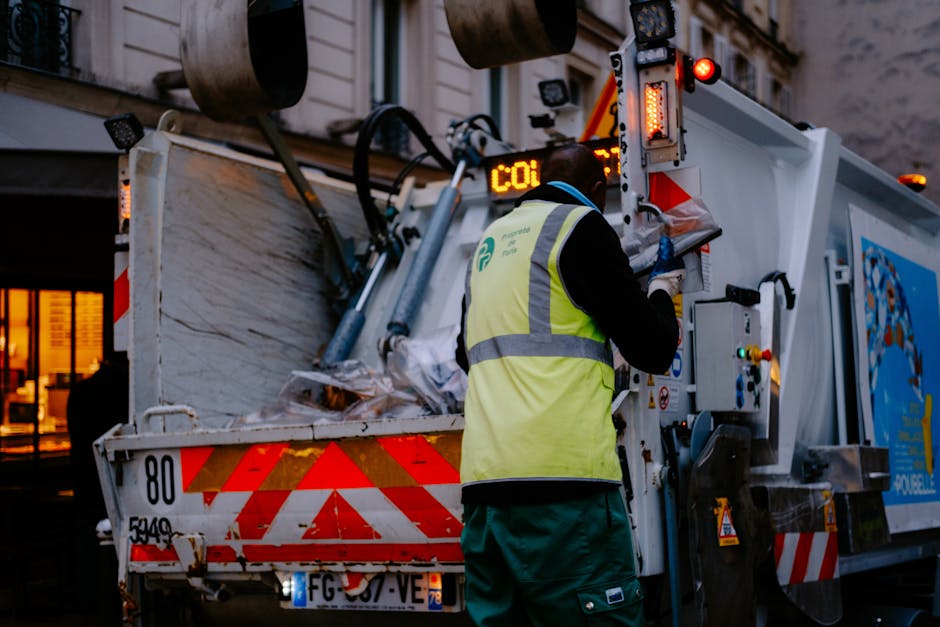 A sanitation worker wearing a high-visibility yellow vest with reflective stripes and dark clothing is operating the controls of a white refuse collection truck parked on a city street during dusk or early evening. The truck's rear compartment is open, revealing a load of mixed waste materials, including cardboard, plastic, and paper, piled inside. The truck has red and orange reflective safety markings on the back, with a license plate partially visible. The worker is focused on managing the collection process, indicating an alternative approach to authorized rubbish disposal, perhaps involving private waste handling services such as those offered by Rubbish Clearance Kensington. In the background, faint outlines of buildings with warm interior lighting suggest an urban environment. The lighting creates a professional, neutral tone suitable for a rubbish removal services context, emphasizing the practicality of on-site waste collection methods.