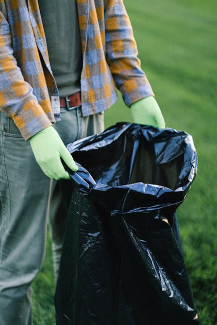 A person wearing a green long-sleeved shirt and a yellow and gray checkered jacket is holding a large black rubbish bag open, preparing for waste collection outdoors. The individual is standing on a grassy area with vibrant green grass in the background, suggesting a park or garden setting. They are wearing light green disposable gloves to handle the rubbish bag safely. The black plastic bag appears to be partially filled, with its interior visible, and is made of shiny, durable plastic material. The scene emphasizes proper waste handling, possibly related to private waste disposal or rubbish removal services provided by companies like Rubbish Clearance Kensington, which offers on-site clearance solutions. The natural outdoor lighting highlights the textures of the clothing, gloves, and the trash bag, creating a clear and detailed visual of the rubbish collection process that aligns with the context of alternative waste management methods beyond local authority services.