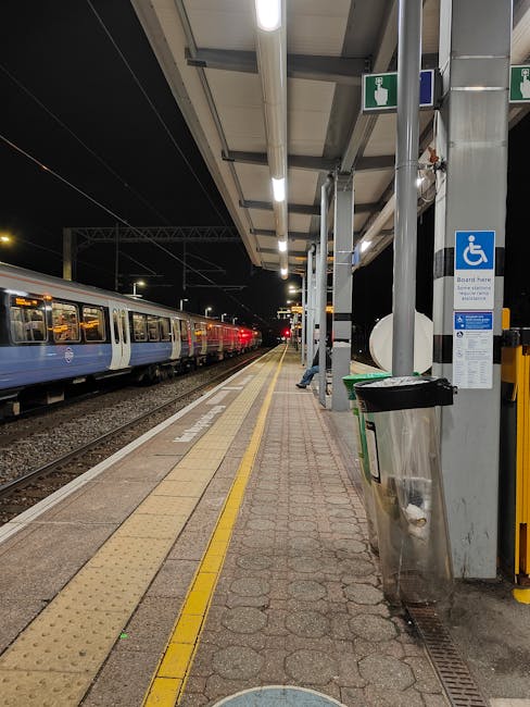 A nighttime scene at South Kensington station platform featuring a row of parked train carriages on the left with blue and white exterior livery, illuminated by overhead lighting fixtures. The platform surface is covered in textured paving stones, with a tactile yellow warning strip running parallel to the tracks. To the right, there is a sheltered waiting area supported by metal poles, with a blue disabled access sign attached to one of the poles. Near the sign, a stainless steel trash bin with a black lid is positioned next to a green box or container, possibly for waste or recycling. The platform appears clean and well-maintained, with minimal clutter, and the environment is quiet, suggesting late evening or night hours. This image from Rubbish Clearance Kensington depicts an urban railway station designed for passenger access, where private waste disposal or rubbish removal services could be relevant for station management or nearby properties, aligning with the context of alternative waste handling and access tips for station environments.