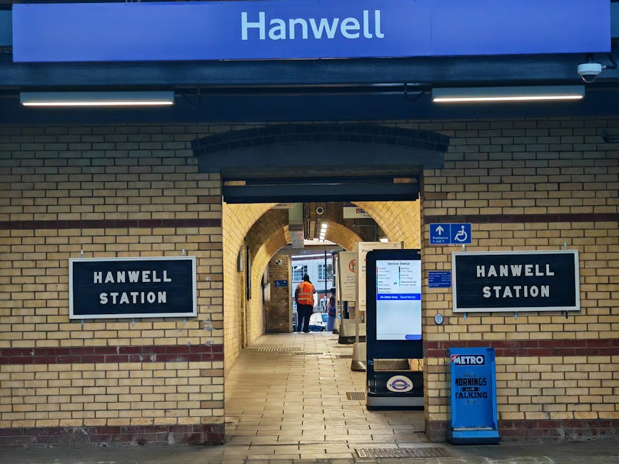 The image depicts the exterior entrance of Hanwell Station, featuring a brick facade with a large blue sign displaying the station's name at the top. Flanking the entrance are two black-framed signs reading 'HANWELL STATION' in white lettering, positioned on either side of the arched passageway. Just below the signs, a smaller blue wheelchair accessibility sign is visible, indicating accessible access points. In front of the entrance, there is a modern digital information kiosk with a white and blue interface, placed on the paved pavement. To the right of the kiosk, a blue Metro-branded bag, likely used for maintenance or waste collection, is situated on the sidewalk. The scene is illuminated by natural daylight, with some artificial lighting above the station signs. Through the arched walkway, a person wearing an orange high-visibility jacket can be seen standing inside, with the station's interior visible beyond, including ticket barriers and additional signage. The environment appears to be clean and orderly, reflecting the typical setting for a suburban railway station in the UK, where independent access and possibly private waste handling are supported by surrounding facilities and services, such as rubbish clearance provided by local providers like Rubbish Clearance Kensington.