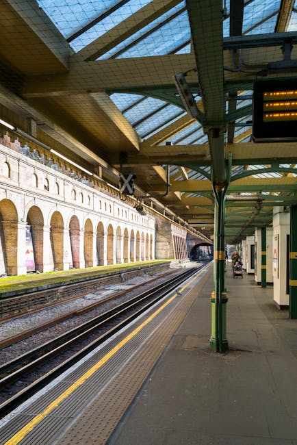 A nighttime scene at South Kensington station platform featuring a row of parked train carriages on the left with blue and white exterior livery, illuminated by overhead lighting fixtures. The platform surface is covered in textured paving stones, with a tactile yellow warning strip running parallel to the tracks. To the right, there is a sheltered waiting area supported by metal poles, with a blue disabled access sign attached to one of the poles. Near the sign, a stainless steel trash bin with a black lid is positioned next to a green box or container, possibly for waste or recycling. The platform appears clean and well-maintained, with minimal clutter, and the environment is quiet, suggesting late evening or night hours. This image from Rubbish Clearance Kensington depicts an urban railway station designed for passenger access, where private waste disposal or rubbish removal services could be relevant for station management or nearby properties, aligning with the context of alternative waste handling and access tips for station environments.